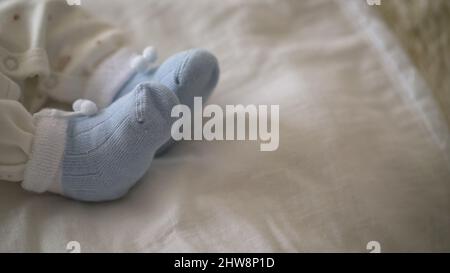 Kleine Beine des Babys liegen auf einer weichen Matratze, tragen Socken. Nahaufnahme eines kleinen Kindes in einem häuslichen Interieur. Babykleidung Konzept. Neugeborenes Baby in blauer Socke Stockfoto