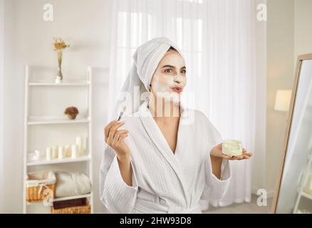 Eine fröhliche Frau legt eine feuchtigkeitsspendende Maske auf ihr Gesicht und hat Spaß bei der Schönheitspflege zu Hause. Stockfoto