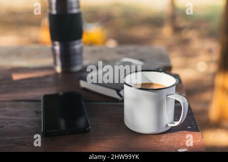 tasse Kaffee auf dem Tisch mit dem Handy und einer Thermoskanne, in einer natürlichen Umgebung Stockfoto