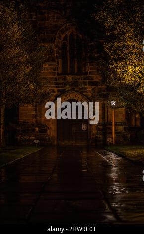 Symmetrische Aufnahme der Prestbury Church, mit dem warmen Licht, das auf dem Steinmaterial der Kirche spielt, und dem nassen Pflaster, das das Licht reflektiert. Stockfoto