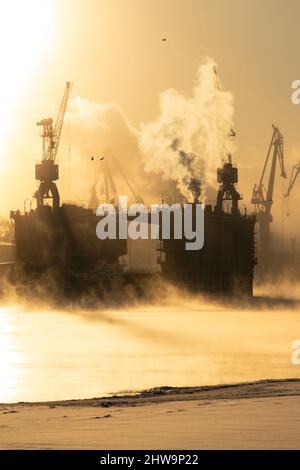 Kränen der Werft in frostigen Wintertag, Dampf über dem Fluss, glatte Oberfläche des Flusses bei Sonnenuntergang. Stockfoto