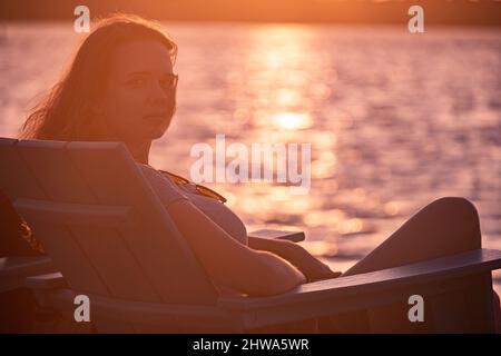 Die junge Frau sitzt am Strand und genießt den Sonnenuntergang über dem Meer Stockfoto