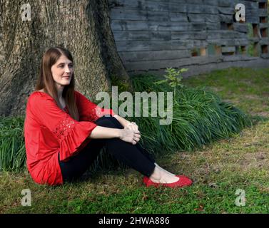 Die junge Frau sitzt auf dem Gras, trägt schwarze Jeans, rote Bluse und Schuhe. Sie schaut in die Ferne und denkt nach. Sie sitzt neben einem Stockfoto