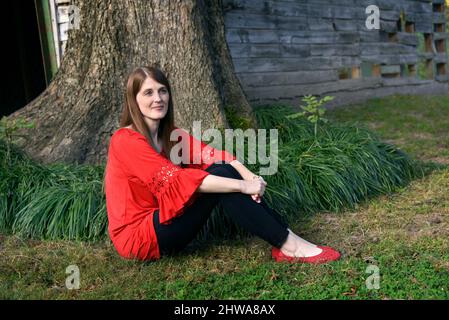 Die junge Frau entspannt sich neben einer rustikalen, hölzernen Scheune. Sie sitzt auf dem Gras neben einem Baum. Ihre Gedanken sind weit entfernt und gelassen. Sie trägt Stockfoto