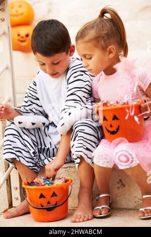 Teilen der Beute. Aufnahme von zwei niedlichen Kindern, die ihre Halloween-Süßigkeiten auscheckten. Stockfoto