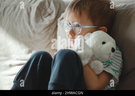 Ein Kind mit Autismus in einer Brille sitzt auf dem Sofa und ist traurig, umarmt sich Stockfoto