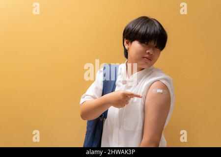 Asiatisch glückliches Mädchen Zeigefinger an Schulter und zeigt ihren Arm mit Verband, nachdem sie in Studentenuniform geimpft wurde, Impfungen für Teenager, Covid preve Stockfoto