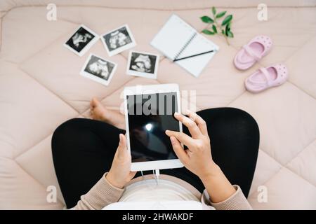 Eine Schwangerschaftsmilcherei. Draufsicht auf die Schwangere, die Gedanken mit einem Tablet für das Gedächtnis niederschreibt, Röntgenbild ihres Babys und ihrer Schuhe.Konzept der Schwangerschaft, Mat Stockfoto