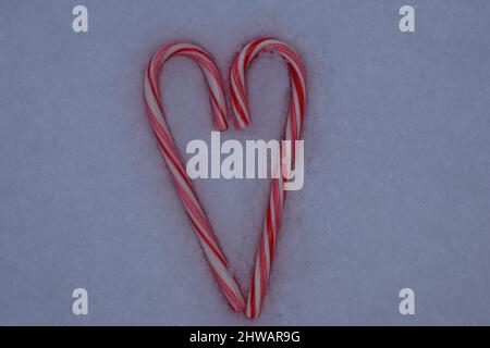 Zwei rot-weiß gestreifte Zuckerstöcke in Form eines Herzens auf Schnee. Stockfoto