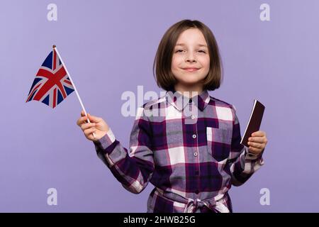 Kind Mädchen 9-11 Jahre alt in Karabinerkleid mit Blick auf die Kamera. Schulmädchen mit Pass und UK-Flagge auf lila Hintergrund, Kopierer Raum, Einwanderung..Study Abroad Konzept. Stockfoto