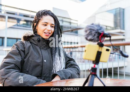 Positive hispanische weibliche Influencerin in warmer Kleidung, die mit dem Smartphone Videos erstellt, während sie am Tisch auf der Terrasse sitzt Stockfoto