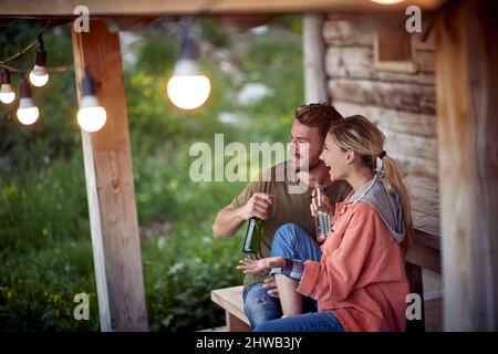 Ein junges, fröhliches Paar, das vor einem Holzhäuschen auf der Terrasse ein Gespräch führt und Bier trinkt. Sommer Gartenfest und Spaß. Freund Stockfoto