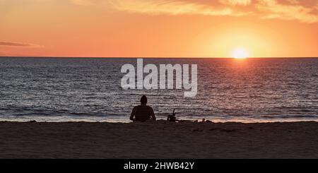 Silhouette eines Mannes, der allein an einem südkalifornischen Strand sitzt und einen strahlenden orangefarbenen Sonnenuntergang mit einem dunklen sandigen Vordergrund beobachtet Stockfoto