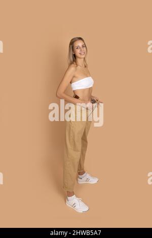 Junge blonde Frau in guter Form lächelnd trägt große übergroße Hosen, um Gewichtsverlust auf hautfarbenem Hintergrund zu zeigen. Halten Sie eine gesunde Ernährung, Bewegung Stockfoto