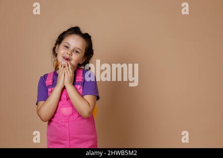 Kind niedlich, kleines Mädchen mit Kanekalon Pigtails der gelben Farbe, berühren Kinn mit Händen Blick auf die Kamera mit sanftem Lächeln trägt rosa Jumpsuit und Stockfoto