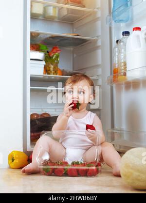 Kann nicht reden. Erdbeeren essen. Aufnahme eines Kleinkindes, das Lebensmittel aus dem Kühlschrank isst. Stockfoto