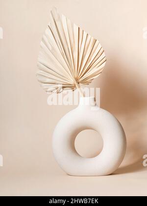 Runde stilvolle Keramikvase mit getrocknetem Palmblatt, das Schatten an die Wand wirft. Beigefarbener Hintergrund. Vorderansicht. Speicherplatz kopieren Stockfoto