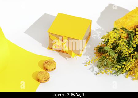 Zweige gelber Mimosenblüten, französischer gelber Makronen und gelber Geschenkkarton mit Schleife. Gruß zum Internationalen Frauentag am 8. März, Motte Stockfoto