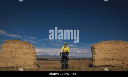 Der Mann reist auf gemischtem Terrain mit Bikepacking. Die Reise des Reisenden mit Fahrradtaschen. Sport Bikepacking, Fahrrad, Sportbekleidung in grün-schwarzen Farben. Schneebedeckter Bergbogen. Stockfoto