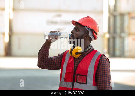 afroamerikanischer Arbeiter, der Wasser trinkt und sich nach der Arbeit auf dem Containerhof ausruht. Stockfoto