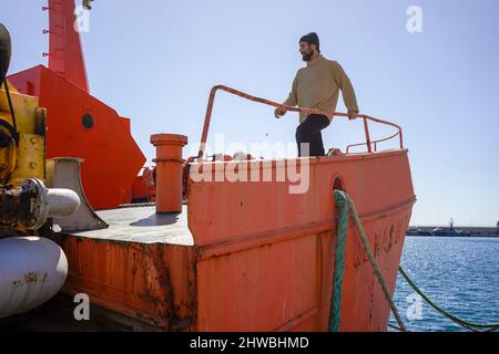 Ein junger, fleißiger Mann mit Bart und Seemann-Pullover erledigt die Arbeit auf den Booten in einem Seehafen Stockfoto