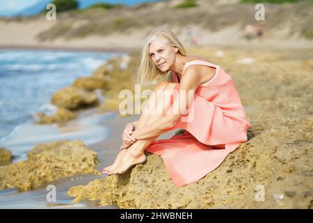 Reife Frau, die auf einigen Felsen am Ufer eines tropischen Strandes sitzt und ein schönes orangefarbenes Kleid trägt. Stockfoto
