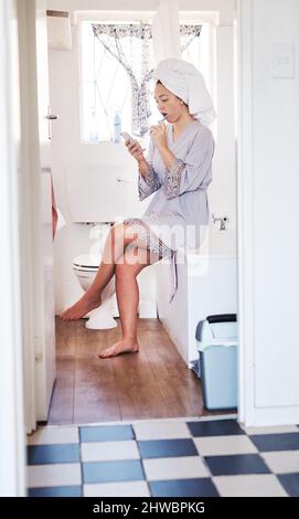 Senden eines Kurztextes während der Auffrischung. Aufnahme einer jungen Frau, die ihr Handy benutzte, während sie sich im Badezimmer die Zähne putzte. Stockfoto