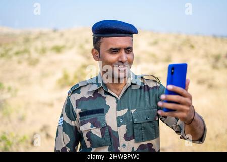 Lächelnde indische Soldaten in Uniform, die während des Dienstes Videoanrufe auf dem Mobiltelefon machen - Konzept der Technologie, fehlende Familie und Entspannung Stockfoto