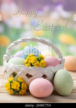 Bunte ostereier mit Korb und Blumen auf einem Holztisch mit buntem Frühlingshintergrund und Happy Easter Text Stockfoto