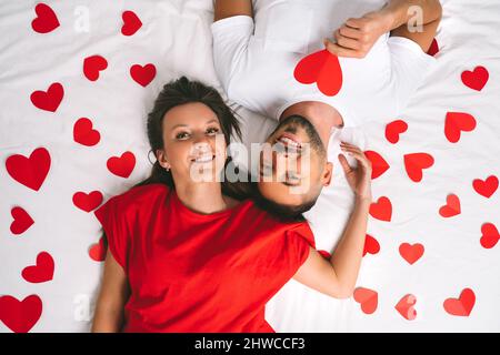 Draufsicht auf ein attraktives verliebtes Paar, das mit roten Papierherzen auf dem Bett liegt Stockfoto