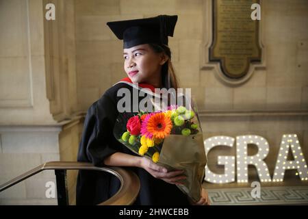 Eine hübsche asiatische weibliche Absolventin mit einem Blumenstrauß posiert für Fotos bei ihrer Abschlussfeier. Dieses formelle Ereignis, bei dem die Graduierung (b Stockfoto