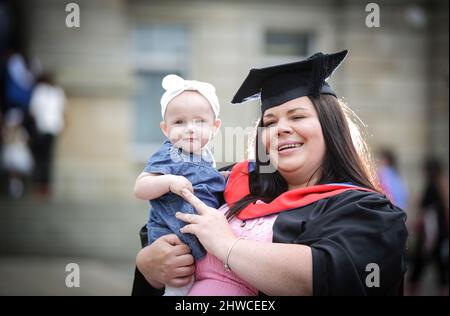 Mutter hält ihre kleine Tochter an ihrem Abschlusstag, versammeln sich die Absolventen, um an der Zeremonie des Abschlusstages teilzunehmen. Dieses formelle Ereignis war graduell Stockfoto