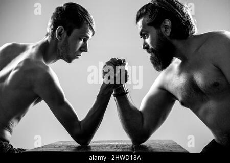 Stark muskulös bärtiger Mann, der einen schwach schwachen Mann am Arm kämpft. Arme ringen dünne Hand, großer starker Arm im Studio. Zwei Männerhände umklammten Arm-Wrestling Stockfoto