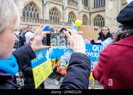 Bath, Großbritannien. 5.. März 2022. Eine Frau macht ein Foto von Demonstranten, die während einer Demonstration gegen die russische Invasion in der Ukraine ein proukrainisches Banner vor der Abtei von Bath halten. Die Demonstration wurde organisiert, um den Menschen vor Ort zu ermöglichen, ihre Solidarität mit der Ukraine im Krieg zwischen Russland und der Ukraine zu zeigen und Putins Aktionen zu verurteilen. Quelle: Lynchpics/Alamy Live News Stockfoto