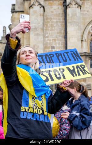 Bath, Großbritannien. 5.. März 2022. Eine Frau, die mit der ukrainischen Flagge drapiert ist, macht während einer Demonstration gegen die russische Invasion in der Ukraine ein Selfie vor der Abtei von Bath. Die Demonstration wurde organisiert, um den Menschen vor Ort zu ermöglichen, ihre Solidarität mit der Ukraine im Krieg zwischen Russland und der Ukraine zu zeigen und Putins Aktionen zu verurteilen. Quelle: Lynchpics/Alamy Live News Stockfoto