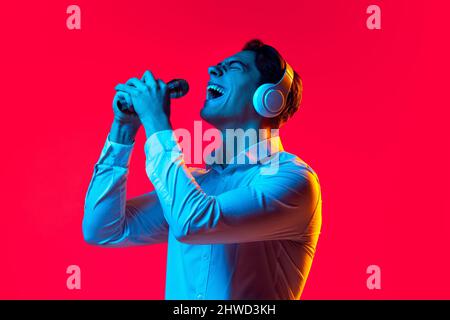 Porträt eines jungen fröhlichen Mannes in weißen Kopfhörern, der im Mikrofon auf rotem Studiohintergrund in blauem Neon singt Stockfoto