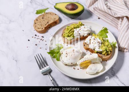 Osterfrühstück oder Brunch. Köstliches Frühstück oder Snack - pochiertes Ei und Frischkäse Toast Vollkorn Roggenbrot, Avocado auf einer Marmorplatte. Stockfoto