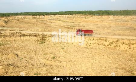 Muldenkipper fährt auf Landstraße. Szene. Draufsicht auf LKW-Fahrten, die vor dem Hintergrund der Baggerkarriere Staubwolken auf unbefestigten Straßen auf dem Land hinterlassen Stockfoto