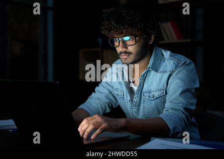 Fokussierter junger indischer Geschäftsmann, der nachts mit einem Laptop online arbeitet. Stockfoto