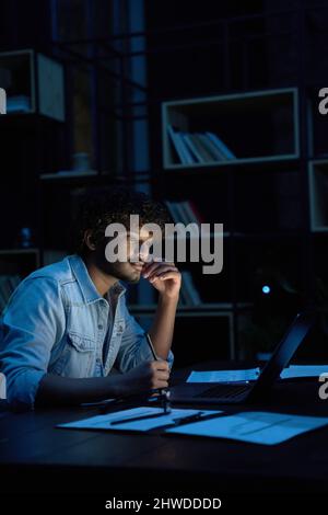Fokussierter junger indischer Geschäftsmann, der nachts mit einem Laptop online arbeitet. Stockfoto
