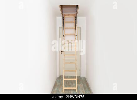 Klappbare Holztreppe im Dachgeschoss auf leerem weißen Hausflur hochziehen. Enger Flur mit weißen Wänden und blauem Laminatboden. Stockfoto
