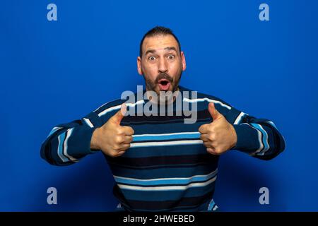 Junger, gutaussehender Mann mit legerer Pullover, der über einem isolierten blauen Erfolgszeichen steht und mit der Hand, dem Daumen nach oben lächelnd und positive Geste macht Stockfoto
