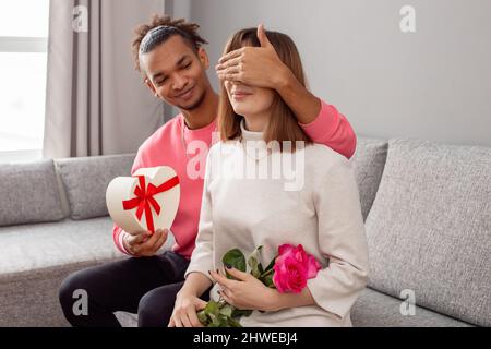 Ein Mann überrascht ein Mädchen, sitzt auf einem grauen Sofa. Stockfoto