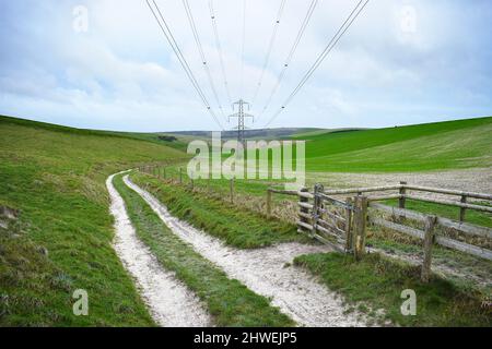 Ein gerutschter Weg neben einem Fußgängertor verläuft in die Distanzen unter elektrischen Kabeln am Himmel mit einem Strommast im Hintergrund. Stockfoto