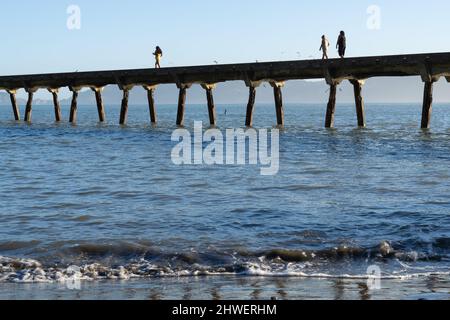 Tokomaru Bay New Zealand - Februar 4 2022; von den auf Meereshöhe ragenden Silhouetten von drei Personen, die auf dem langen historischen Tokomaru Bay Wharf in die Bucht laufen Stockfoto