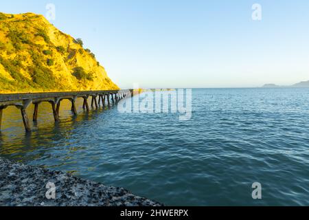 Sonnendurchflutete Klippen und historische Anlegestelle am Ende der Tokomaru Bay. Stockfoto