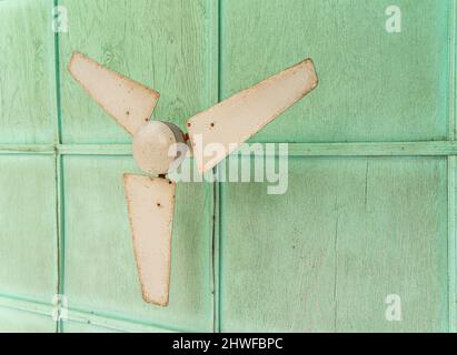 Elektrisch angetriebener staubiger weißer Deckenventilator, der an einer alten, grünen Decke montiert ist Stockfoto