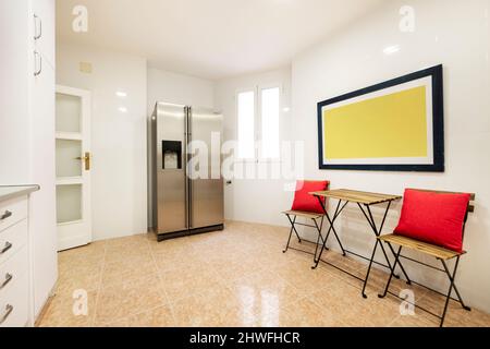 Küche mit einem großen Edelstahl-Kühlschrank und einem kleinen Holztisch mit zwei Klappstühlen an einer Wand. Stockfoto