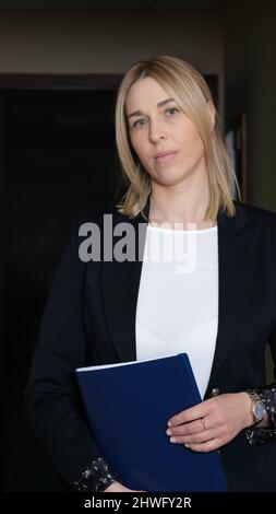Frau in einem schwarzen formalen Blazer hält Dokumente und Blick auf die Kamera. Blonde selbstbewusste Frau Business-Chef stehend mit Ordner in den Händen Stockfoto