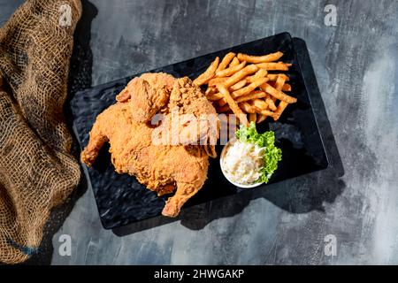 Knusprig gebratenes Spring Chicken (voll) mit pommes Frites und Mayo Dip in ein schwarzes Gericht Draufsicht auf dunklem Hintergrund Fast Food Stockfoto